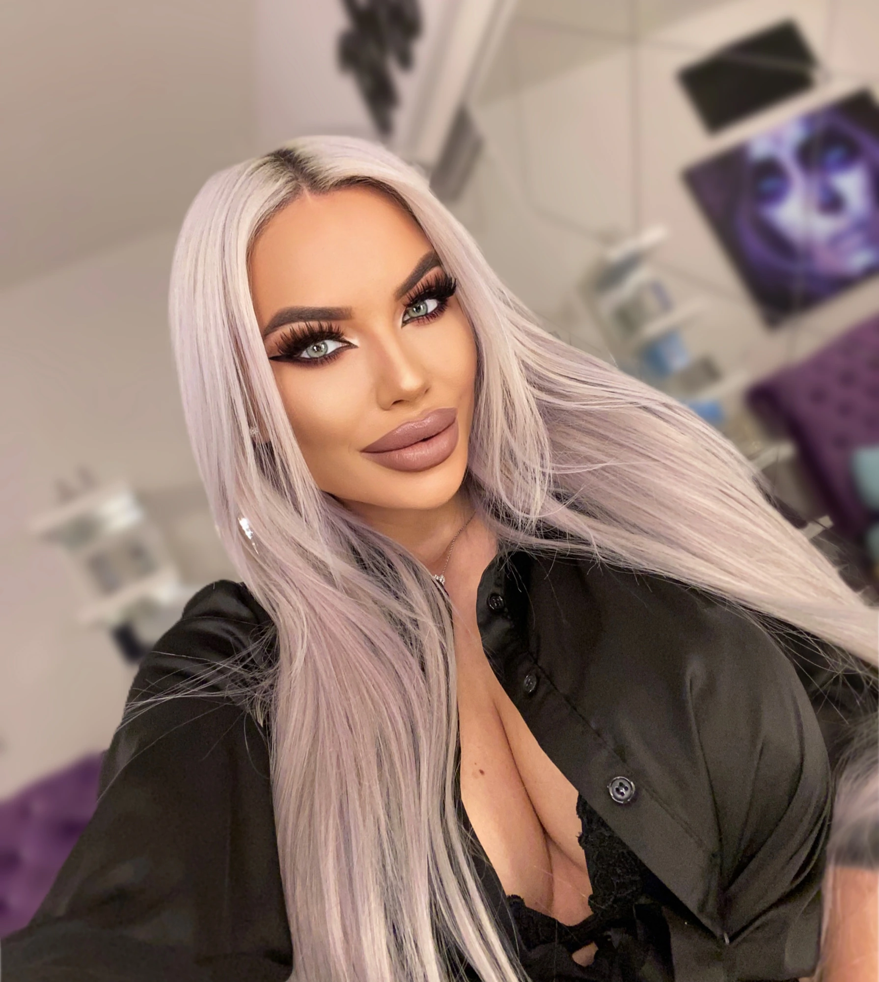 Sasha with long silver-blonde hair in an open black shirt, in a softly lit room with purple velvet behind her.