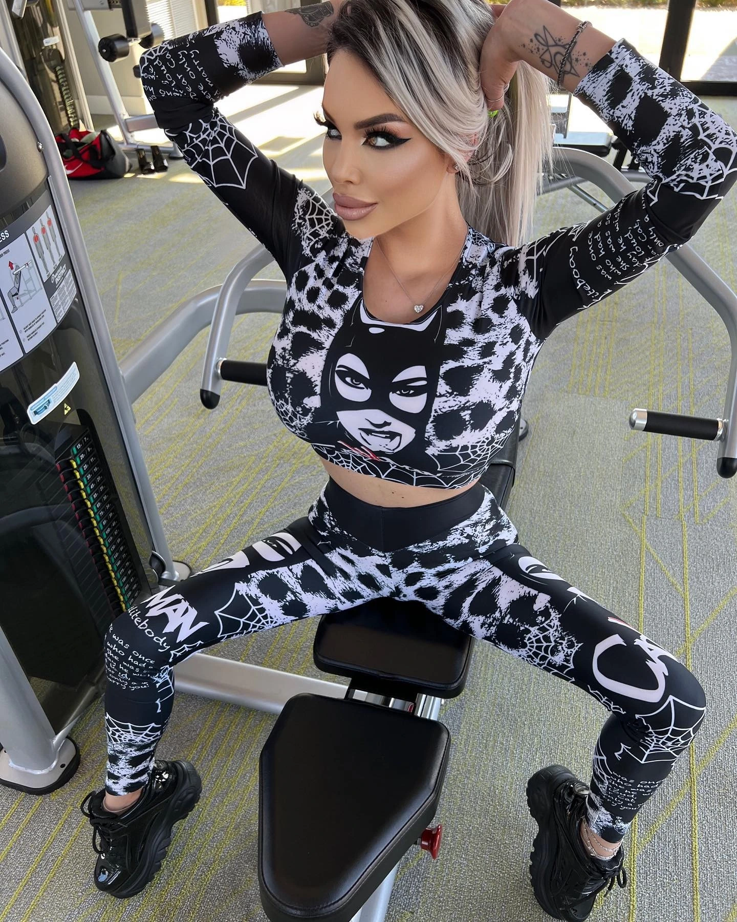 Sasha on a weight machine with her hands behind her head, in the black and white printed crop top and leggings and chunky black sneakers.