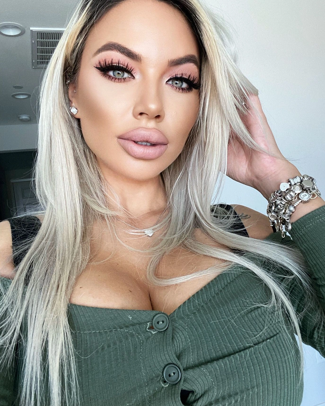 Sasha in an olive ribbed knit, one hand in her hair and a stack of silver charm bracelets on her wrist.
