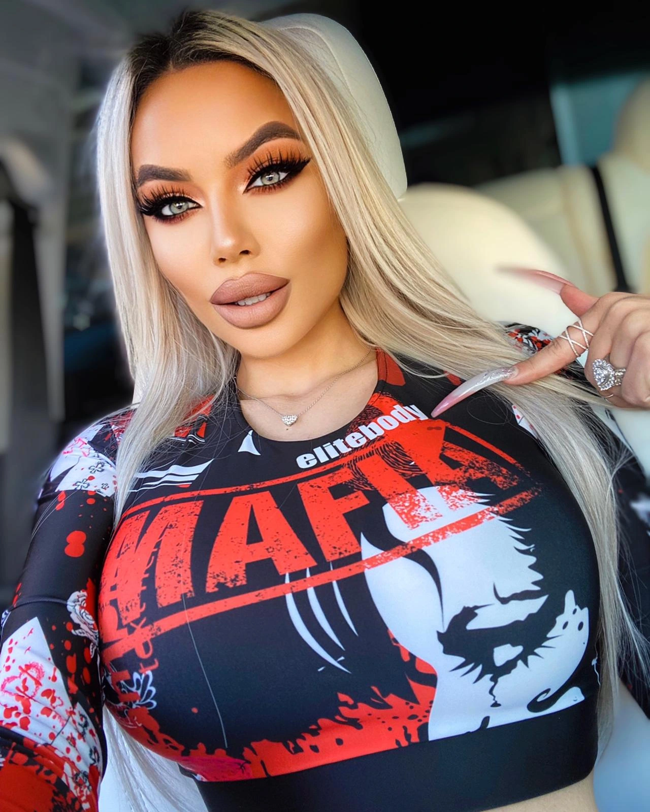 Sasha in the car in a black, red and white elitebody MAFIA graphic crop top, her long platinum hair loose, pointing at the print with long nude nails.