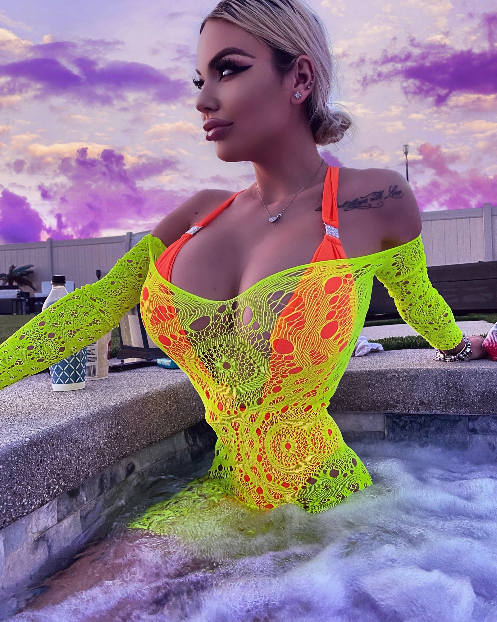 Sasha in a hot tub at sunset in a neon yellow and orange crochet top, her hair in a low bun, under a pink and purple sky.