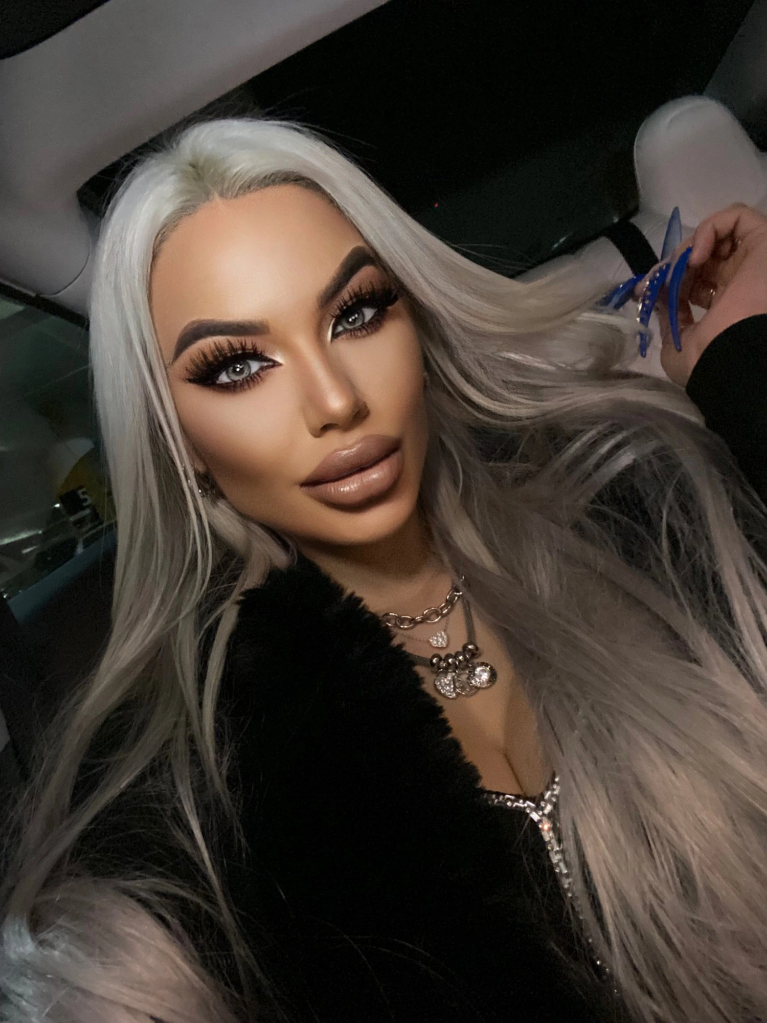 A close-up of Sasha at night in the car: platinum hair, soft bronze glam and a glossy nude lip, layered silver chains over black fur.