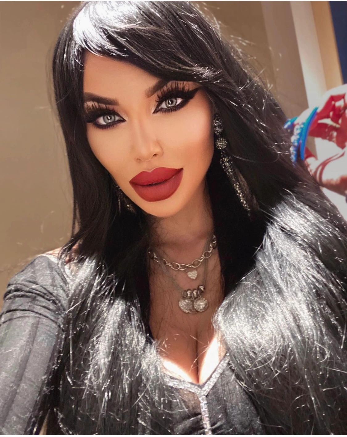 Sasha with long black hair, a matte red lip and dramatic winged liner, in crystal drop earrings, layered silver chains and a glittery silver-and-black tinsel top.