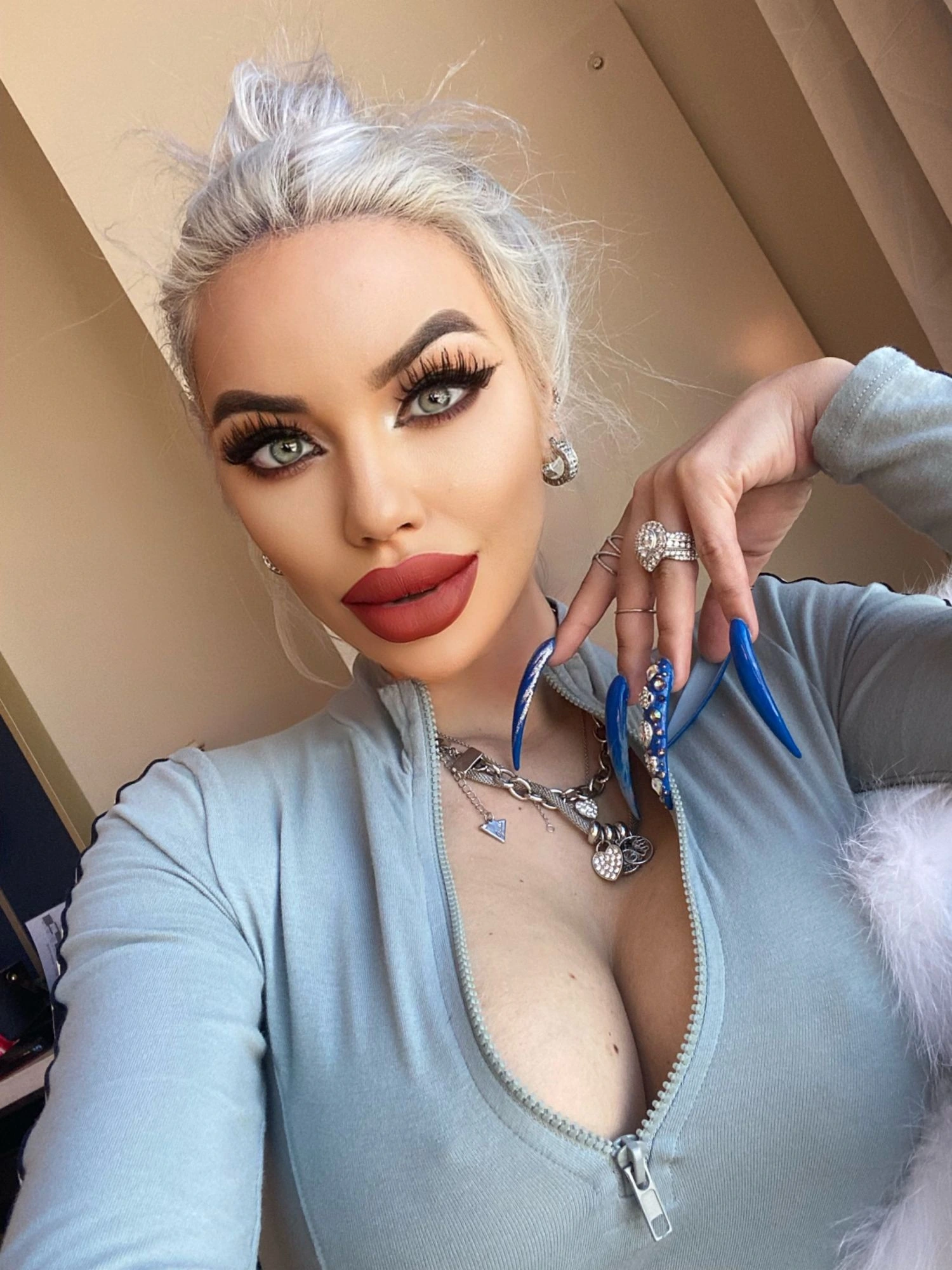 Sasha with a red lip and long blue stiletto nails, her platinum hair pinned up, in a pale blue zip top.