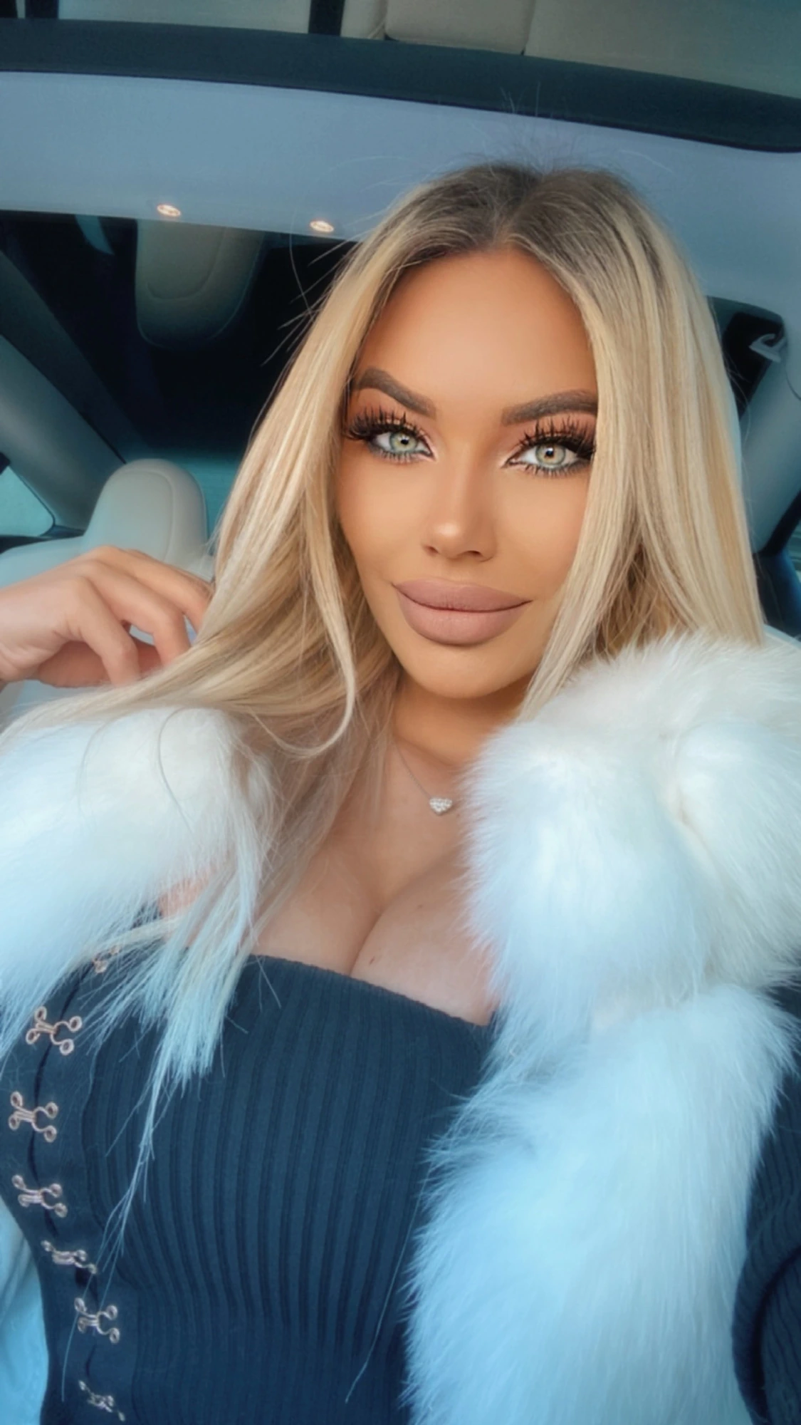 Sasha in the car in a white fur jacket over a navy ribbed top, her long blonde hair loose and one hand raised to it.