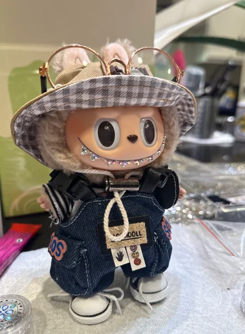 Labubu Nº 06: houndstooth bucket hat with gold glasses, denim overalls and a crystal smile.